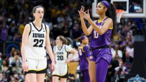 An image of Caitlin Clark and Angel Reese. Angel Reese is wearing her purple LSU uniform while taunting Clark, who is wearing her white Hawkeye's jersey.