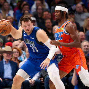 A picture of Luka Doncic in the post wearing the Mavericks blue jerseys, while being defended by Shai Gilgeous-Alexander wearing an orange Oklahoma City Thunder jersey.